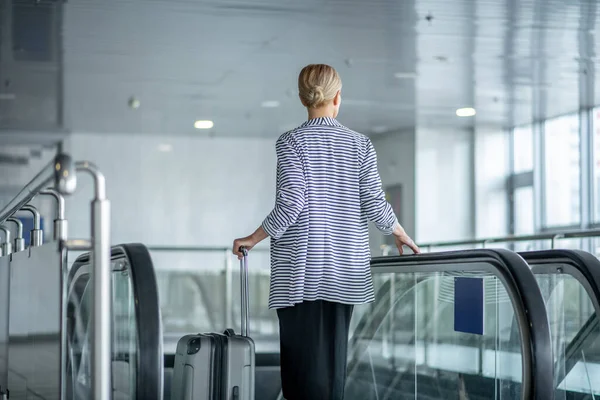 Elegant female tourist with the trolley suitcase riding the escalator — Stock fotografie
