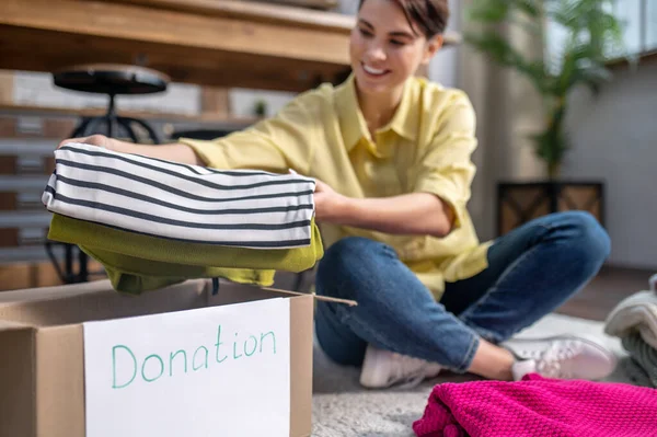 Joyful girl seated on the carpet packing things for donation — Stockfoto