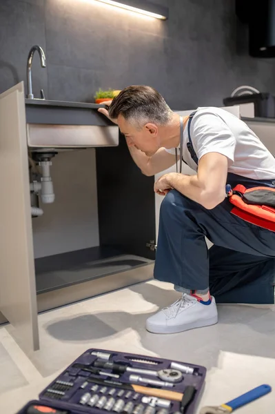 Man kneeling down to inspect pipes under sink — 스톡 사진