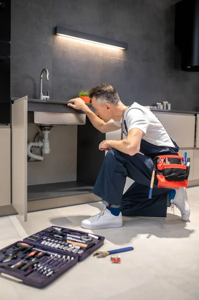 Man kneeling down touching sink inspecting pipes — 图库照片