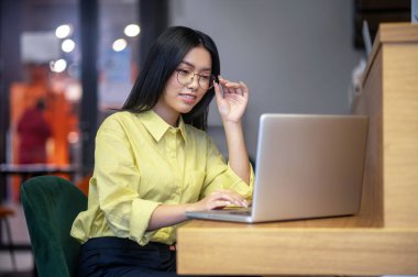 Cute asian young woman in eyeglasses at the laptop having coffee