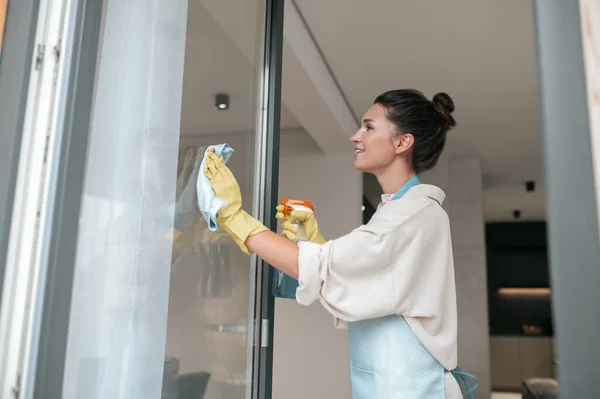 Una mujer en delantal limpiando las ventanas y buscando involucrada —  Fotos de Stock