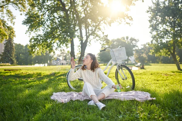 A young girl sitting on the grass and having fun making bubbles — Stock Photo, Image