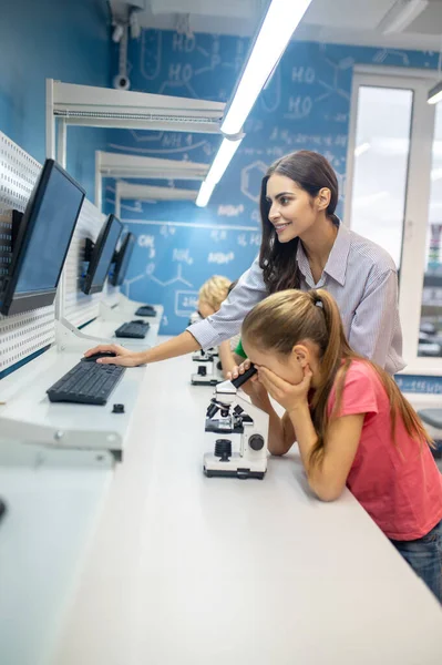 Kobieta patrząc na ekran i dziewczyna pochyla się nad mikroskopem — Zdjęcie stockowe