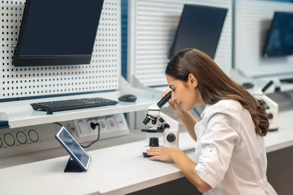 女人坐在桌边透过显微镜看 — 图库照片