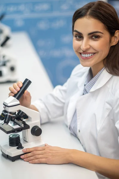 Женщина, касающаяся микроскопа, улыбается в камеру — стоковое фото