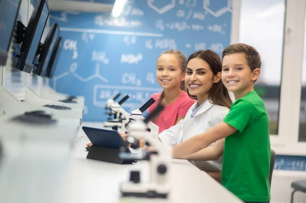 현미경 가까이에 있는 어린이들 이 카메라를 보고 웃고 있는 교사 — 스톡 사진