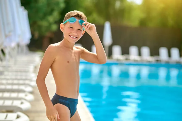 Boy touching goggles smiling at camera near pool — Stock Photo, Image