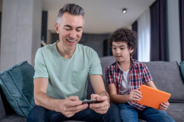 Çocuk yanında oturan adamın akıllı telefonuna bakıyor.
