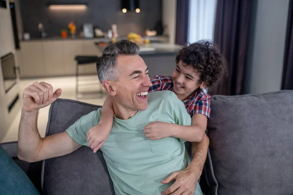 Hijo abrazando riendo padre sentado en sofá —  Fotos de Stock