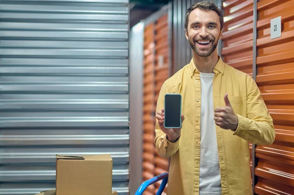 Joyeux jeune ouvrier d'entrepôt montrant son téléphone portable — Photo