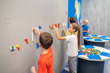 Duvarın yanında duran çocuklar lego mozaiği yapıyorlar.