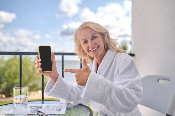 Блондинка в білому халаті зі смартфоном в руках виглядає щасливою — стокове фото