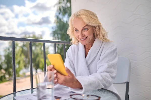 Блондинка в белом халате со смартфоном в руках, выглядящая счастливой — стоковое фото