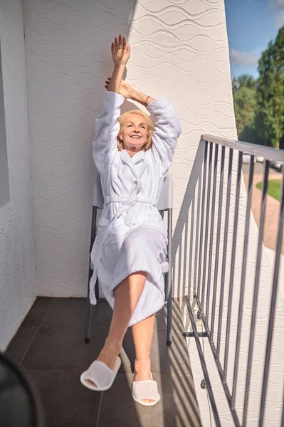Egy szőke középkorú nő fehér köntösben élvezi a napsütést. — Stock Fotó