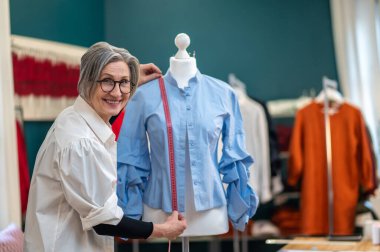 Kameraya gülümseyen bir kadın bluza ölçü alıyor