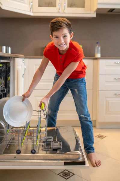 Lächelndes Kind bereitet Geschirr zum Reinigen in der Spülmaschine vor — Stockfoto