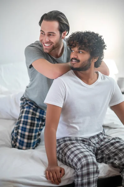 Unga biracial gay par i pyjamas sitter på sängen — Stockfoto