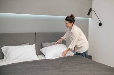 Genç bir kadın yatağını yapıyor ve pozitif görünüyor.