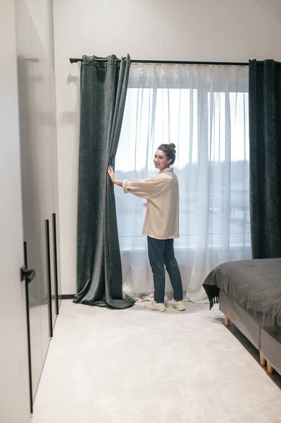 En kvinna med en hårbulle som öppnar gardinerna i rummet — Stockfoto