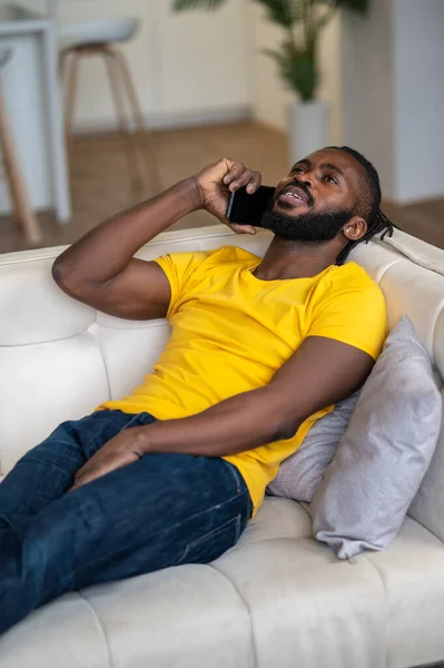 Hombre de piel oscura hablando en un smartphone tumbado en un sofá — Foto de Stock