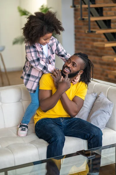 Girl standing on couch hugging her sitting dad — Stock Photo, Image