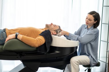 Erkek müşteri güzellik salonundaki sandalyede oturuyor ve saçlarını yıkatıyor.