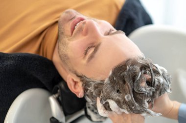 Erkek müşteri güzellik salonundaki sandalyede oturuyor ve saçlarını yıkatıyor.