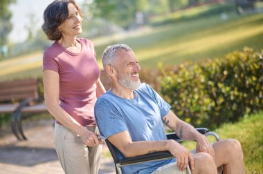 Siyah saçlı bir kadın kocasını tekerlekli sandalyede taşıyor.