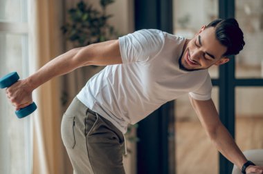 Koyu renk saçlı genç adam halterlerle egzersiz yapıyor ve halinden memnun görünüyor.