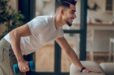 Koyu renk saçlı genç adam halterlerle egzersiz yapıyor ve halinden memnun görünüyor.