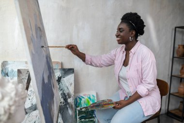 Esmer tenli bir kadın şövalenin yanında oturuyor ve resim yapıyor.