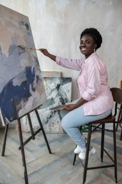 Esmer tenli bir kadın şövalenin yanında oturuyor ve resim yapıyor.