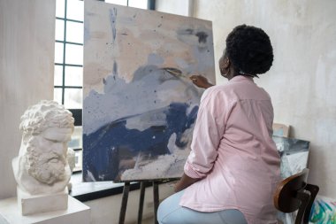 Esmer tenli bir kadın şövalenin yanında oturuyor ve resim yapıyor.