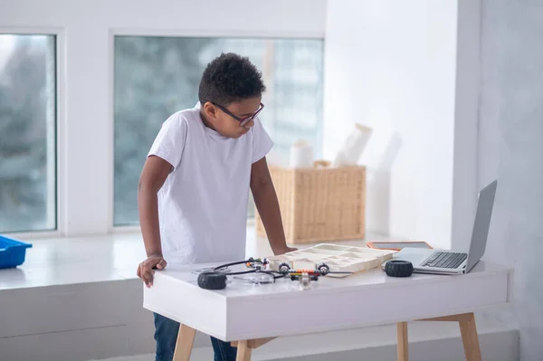 Un niño con una camiseta blanca de pie en la mesa con cables y portátil —  Fotos de Stock