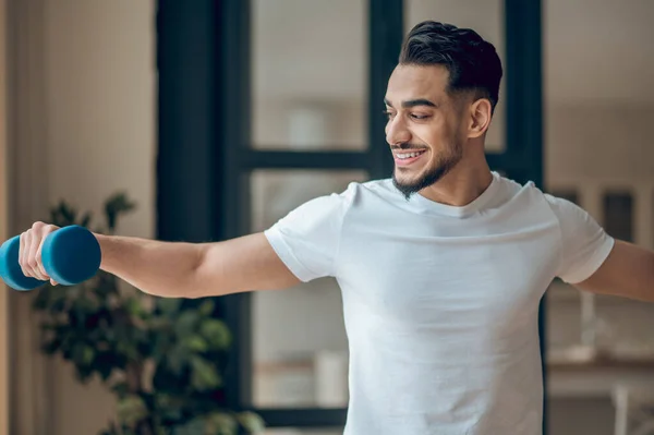 Jovem homem de cabelos escuros se exercitando com halteres e olhando satisfeito — Fotografia de Stock