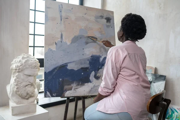 Esmer tenli bir kadın şövalenin yanında oturuyor ve resim yapıyor.