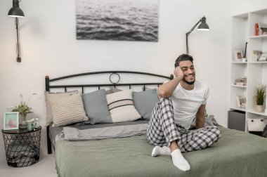 Beyaz tişörtlü ve ekose pantolonlu bir adam evde yatakta oturuyor.