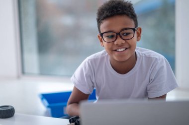 A smiling dark-skinned boy sitting at the laptop clipart