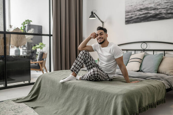 A man in white tshirt and plaid pants sitting on bed at home