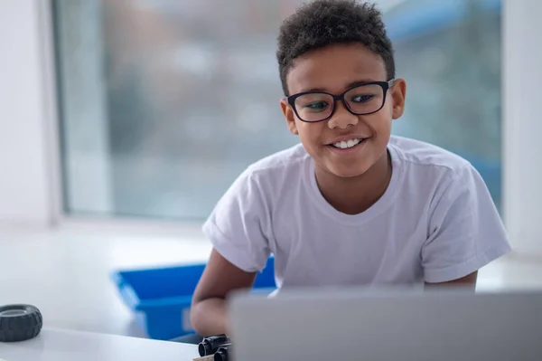 Egy mosolygó, sötét bőrű fiú ül a laptopnál. — Stock Fotó