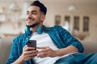 Mavi tişörtlü, elinde telefonla kanepede oturan genç adam.