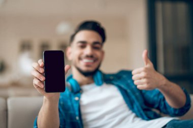 Mavi tişörtlü, elinde telefonla kanepede oturan genç adam.