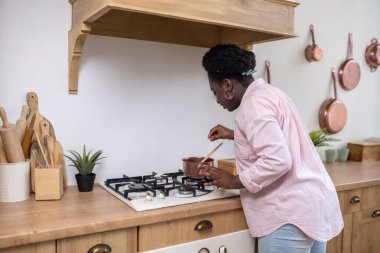 Artı, mutfakta yemek pişiren pembe elbiseli Afrikalı kadın.