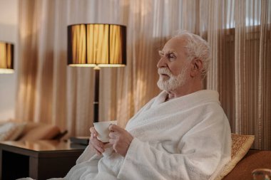 Gri saçlı son sınıf öğrencisi sabah kahvesini içiyor ve düşünceli görünüyor.