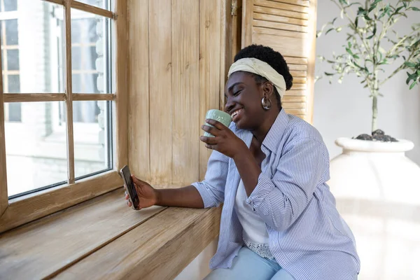 窓の近くにスマートフォンを持っているアフリカの女性 — ストック写真