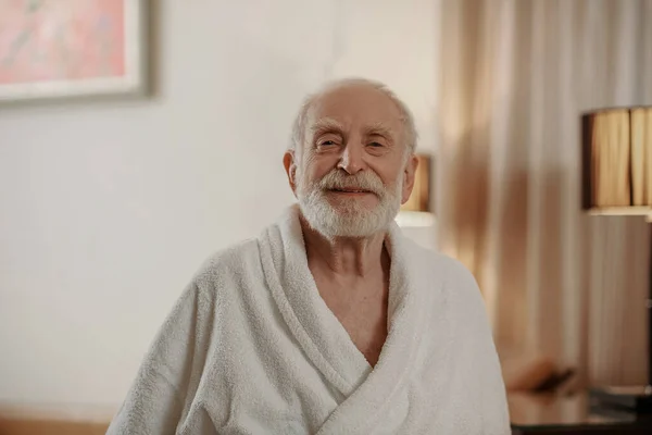 A gray-haired bearded man in a white robe in a hotel room — стоковое фото