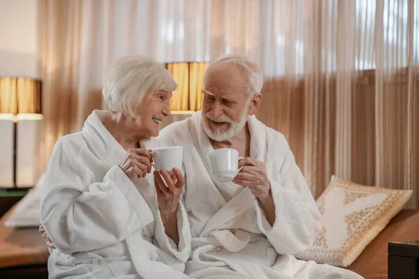 A senior couple in white robes having coffee and talking — стоковое фото