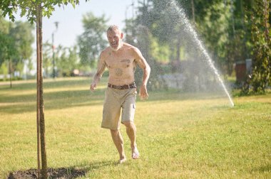 Parkta duşun altında şortlu yarı çıplak bir adam.