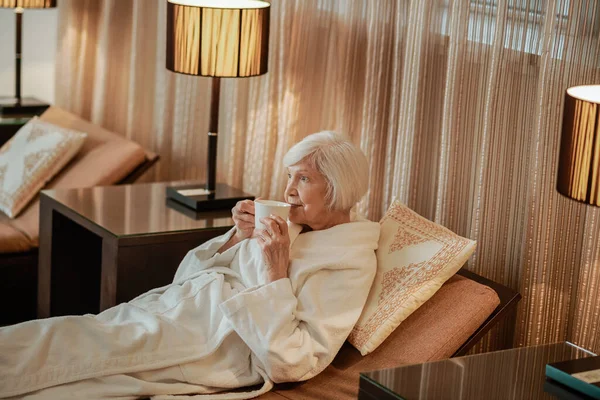A senior gray-haired woman lying in bed and having morning coffee — Fotografia de Stock